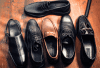 Six pairs of men's dress shoes in various styles, including loafers, oxfords, and brogues, arranged on a wooden surface. The shoes are in black and brown shades, with some featuring decorative details like perforations and metallic accents.