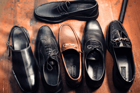 Six pairs of men's dress shoes in various styles, including loafers, oxfords, and brogues, arranged on a wooden surface. The shoes are in black and brown shades, with some featuring decorative details like perforations and metallic accents.