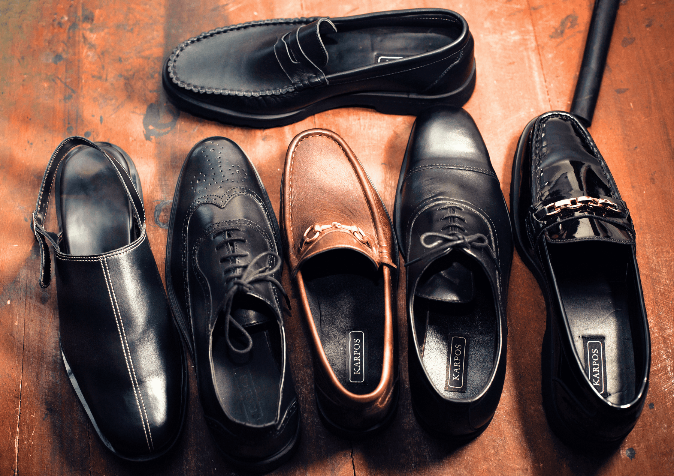 Six pairs of men's dress shoes in various styles, including loafers, oxfords, and brogues, arranged on a wooden surface. The shoes are in black and brown shades, with some featuring decorative details like perforations and metallic accents.